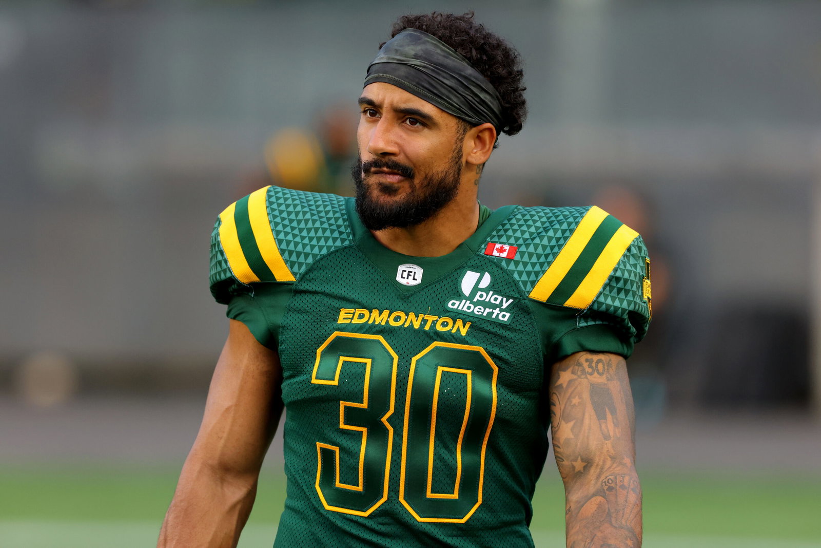A football player wearing a green and yellow Edmonton jersey with the number 30, featuring a headband and visible tattoos on their arm, stands on a field.