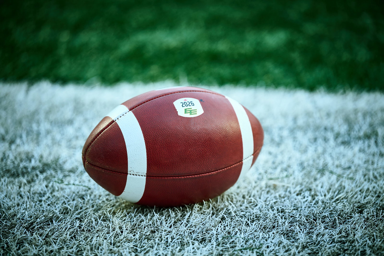 A brown football with white stripes rests on frost-covered grass, featuring a "2026" sticker.