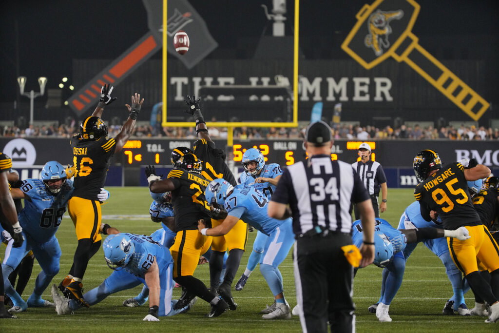 Football players in black and yellow and light blue uniforms compete for a ball near the goalpost during a game.