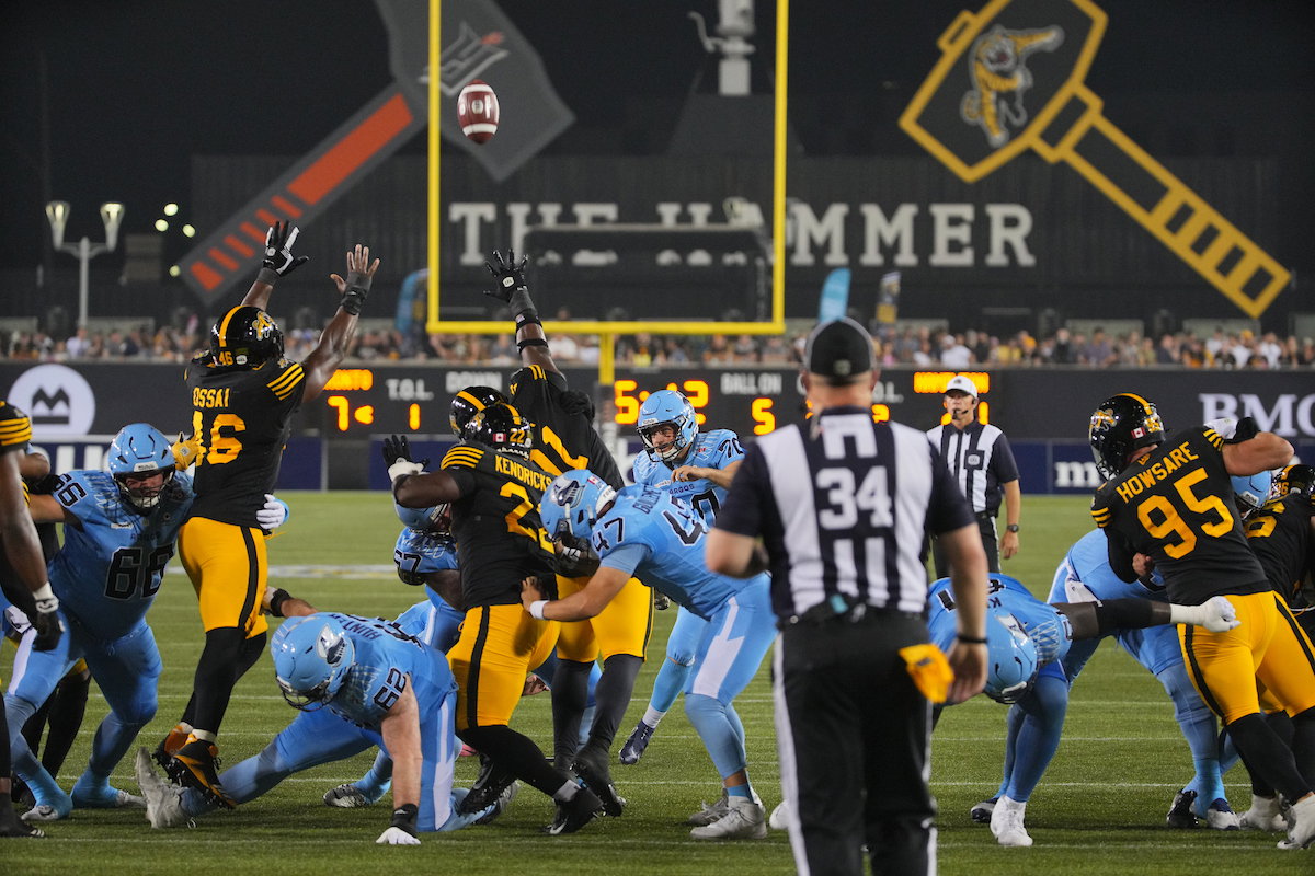 Football players in black and yellow and light blue uniforms compete for a ball near the goalpost during a game.