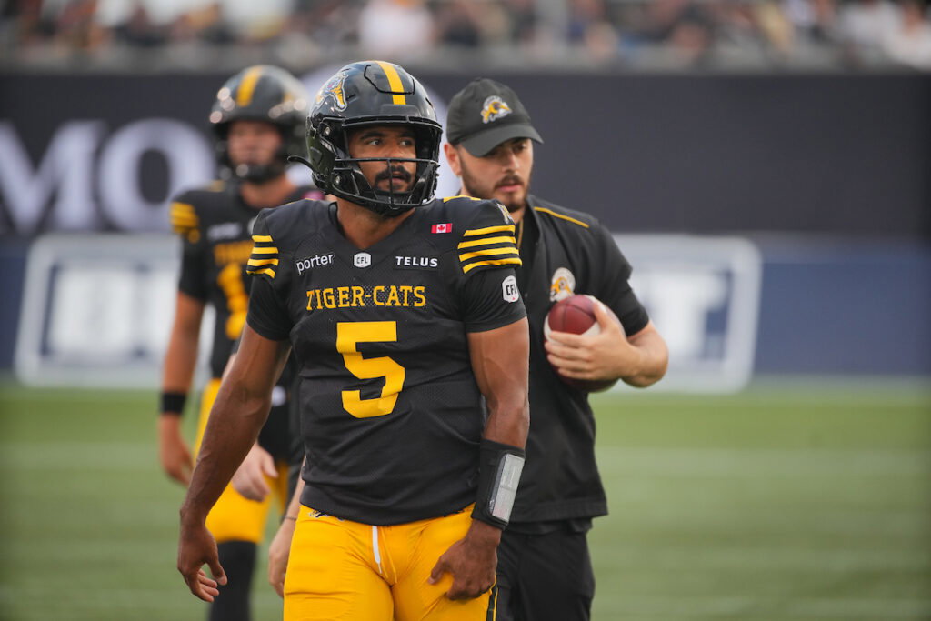 A football player wearing a black and yellow Tiger-Cats jersey with the number 5 walks on the field, accompanied by a coach holding a football.
