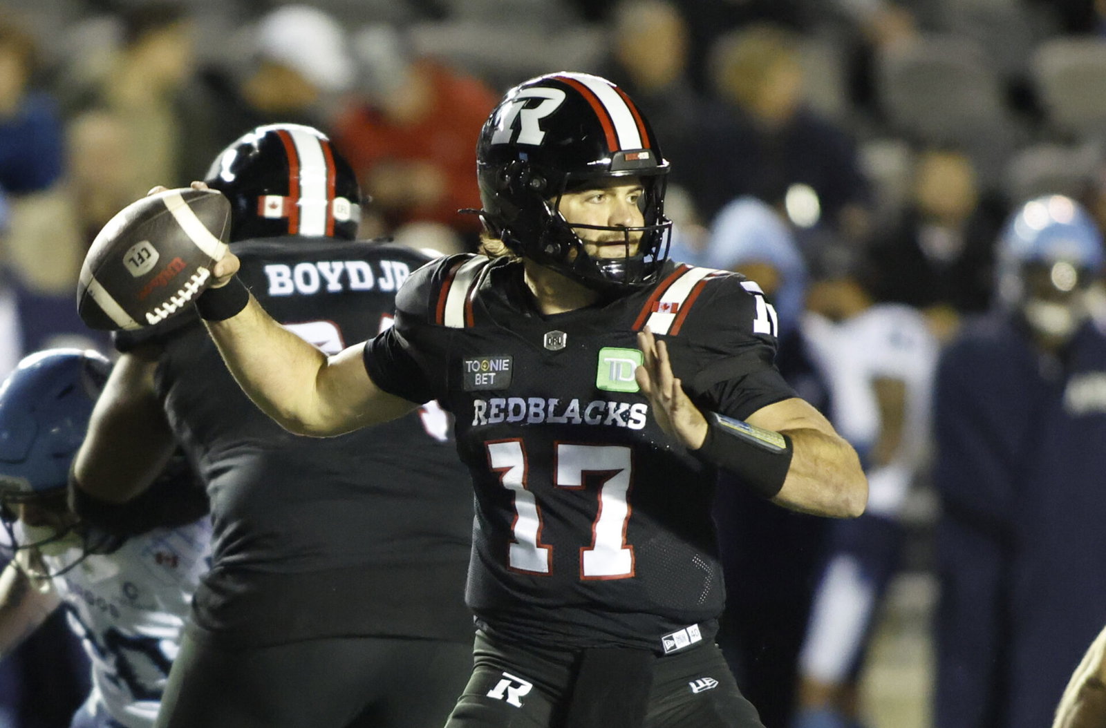 A football player in a black jersey with the number 17 and "REDBLACKS" on the front is preparing to throw the ball during a game.