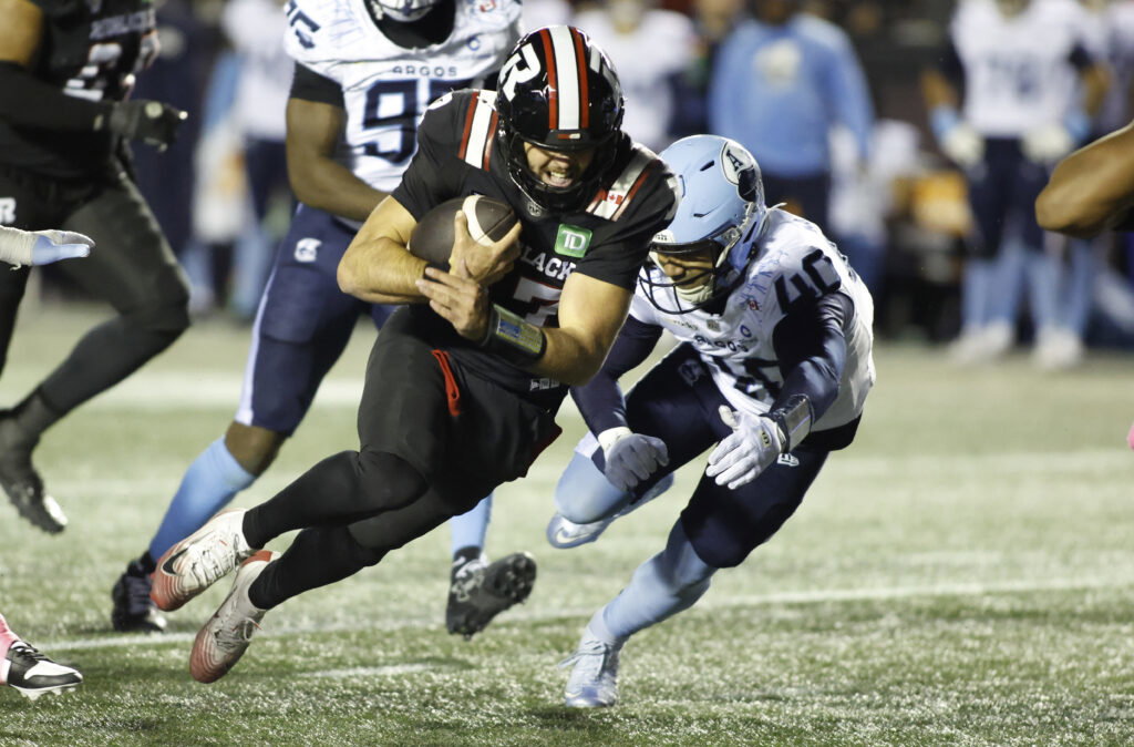 A football player in a black uniform runs with the ball while being pursued by an opponent in a light blue uniform.