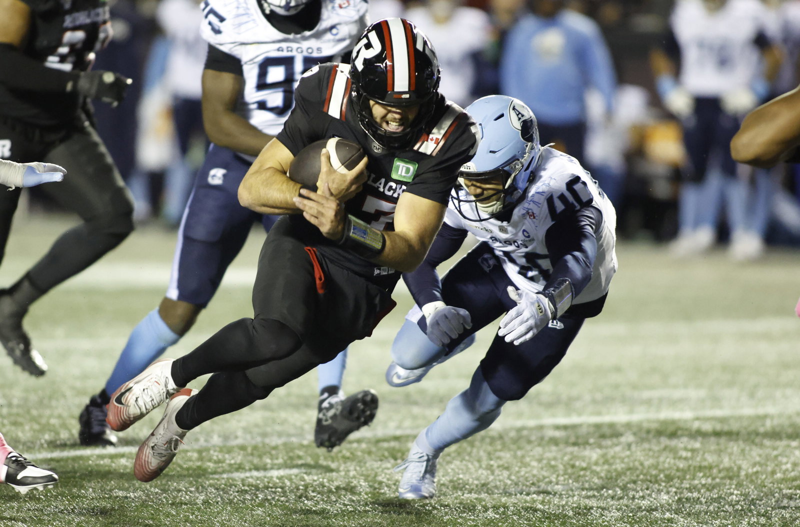 A football player in a black uniform runs with the ball while being pursued by an opponent in a light blue uniform.